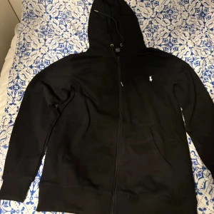 Svart hoodie från Polo Ralph Lauren - Svart hoodie från Polo Ralph Lauren med dragkedja och huva. Klassisk design med vit broderad logga på bröstet, känguruficka och ribbade muddar. Perfekt för chill dagar eller när du vill ha en clean streetstyle-look.