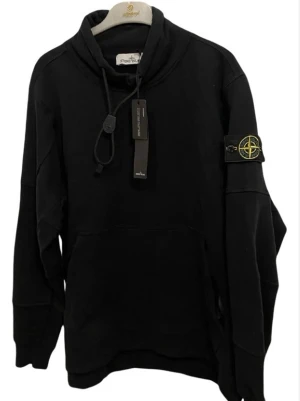 Svart hoodie från Stone Island - Säljer en svart hoodie från Stone Island med klassisk patch på ärmen och justerbar huva med dragsko. Hoodien har en stor magficka och är gjord i mjukt bomullsmaterial. Perfekt för dig som gillar streetwear och vill ha en stilren look.