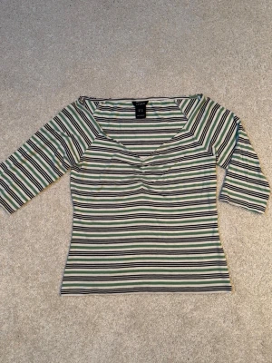 Randig topp med v-ringning från Lindex - Snygg randig topp från Lindex med v-ringad hals och trekvartsärm. Färgerna går i vitt, svart och grönt. Toppen har en lätt åtsittande passform och är gjord i mjukt material. Perfekt för en avslappnad och trendig look. Sparsamt använd, som ny.