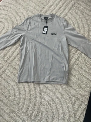 Ljusgrå långärmad topp från EA7 - Snygg ljusgrå långärmad topp från EA7 Emporio Armani med rund halsringning och svart logga på bröstet. Tillverkad i mjukt och lätt material som känns skönt mot huden. Perfekt för dig som gillar stilrena och sportiga plagg.