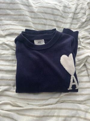 Mörkblå stickad tröja från Ami - Snygg mörkblå stickad tröja från Ami med rund halsringning och ribbade muddar. På bröstet finns ett vitt hjärta och bokstaven A som ger en unik touch. Perfekt för dig som gillar stilrena plagg med en twist.