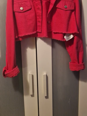 Croppad röd jeansjacka - Snygg croppad röd jeansjacka med rå kant och klassiska bröstfickor. Jackan har långa ärmar och knappar framtill. Perfekt för dig som vill sticka ut med en färgstark och trendig look. På bilden ser den typ röd ut men den är chockrosa. Fins katt i hemmet