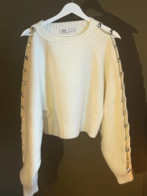 Vit stickad tröja med silverknappar Zara - Vit stickad tröja från Zara med rund halsringning och dekorativa silverfärgade knappar längs hela ärmarna. Tröjan har en rak, något boxig passform och ribbstickade muddar vid ärmslut och hals. Perfekt för dig som gillar unika detaljer. Denna är så fin och det är smärtsamt att sälja den! Köpt från Zara, helt nyskick🤍