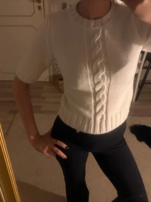 Vit stickad topp med fläta - Säljer en vit stickad topp med korta ärmar och en snygg flätstickad detalj framtill. Tröjan har ribbade muddar vid halsringning och nederkant, och är något croppad i modellen. Perfekt till både jeans och kjol.💕