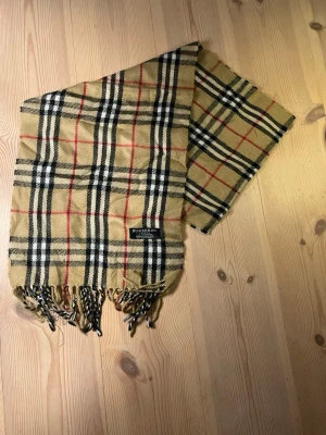 Burberry halsduk i klassiskt rutmönster - Säljer en beige halsduk från Burberry med det ikoniska rutmönstret i svart, vitt och rött. Halsduken har fransar i ändarna och är tillverkad i mjukt material som känns lyxigt och stilrent. Perfekt accessoar för att lyfta din outfit. Pris kan diskuteras.