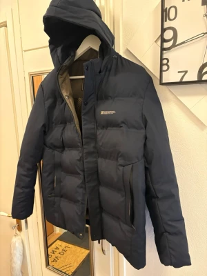 Mörkblå pufferjacka med huva - Säljer en mörkblå pufferjacka från Jingpin med stor huva och två dragkedjeförsedda fickor. Jackan har quiltad design och insidan är fodrad med mönstrat tyg i beige och guld. Perfekt för kalla dagar och har en clean, modern look.