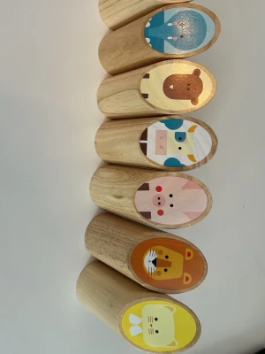 Träklossar med djurmotiv - Sex stycken ovala träklossar med färgglada djurmotiv på ena sidan. Motiven inkluderar blå elefant, brun björn, vit och blå ko, rosa gris, orange lejon och gul katt. Klossarna är tillverkade i ljust trä och har en slät, rundad form som är skön att hålla i.