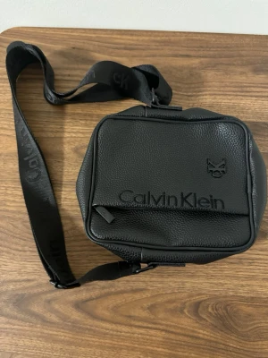 Svart axelväska från Calvin Klein - Snygg svart axelväska från Calvin Klein i strukturerat skinn med justerbar axelrem och logotyp framtill. Väskan har dragkedja både fram och bak samt ett stilrent, minimalistiskt utseende. Perfekt för att bära det viktigaste med stil.