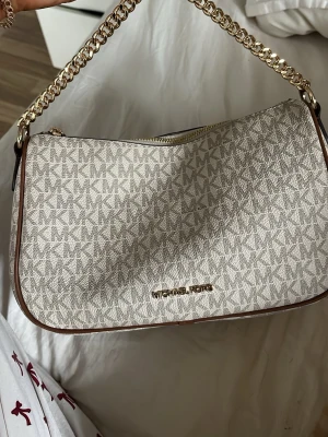 Michael Kors beige handväska - Snygg beige handväska från Michael Kors med kedjerem i guld och klassiskt MK-mönster över hela väskan. Väskan har dragkedja upptill och bruna detaljer längs kanterna. Perfekt storlek för det viktigaste och ger en lyxig känsla. Original pris - 3500kr
