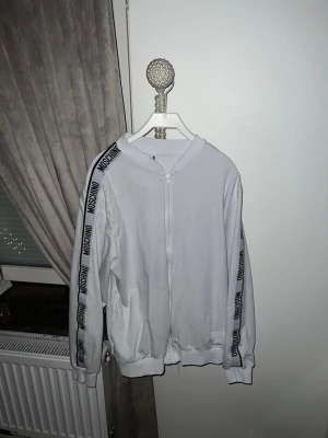Vit ziptröja från Moschino - Säljer en vit ziptröja från Moschino med svarta logotexter längs ärmarna. Tröjan har dragkedja framtill, ribbade muddar och en avslappnad lock, Den är helt äkta och köpt för 3000kr 