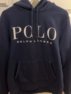 Mörkblå hoodie från Polo Ralph Lauren - Snygg mörkblå hoodie från Polo Ralph Lauren med stor vit logga på bröstet. Klassisk huva med dragsko och en stor magficka framtill. Tillverkad i mjukt bomullsmaterial som är skönt att ha på sig. Perfekt för en avslappnad och stilren look. Hoodien är i perfekt skick. Storlek: M. Nypris: 2000 kr