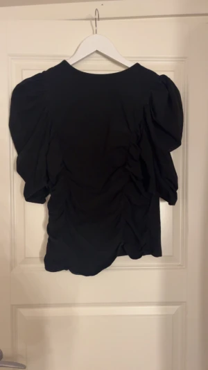 Svart blus med puffärm och rynk - Säljer en svart blus med puffiga ärmar och snygga rynkade detaljer längs sidorna. Blusen har en rund halsringning och är tillverkad i ett mjukt tyg som ger en skön känsla. Perfekt för dig som gillar volym och coola detaljer på toppen. Lite av rynkorna har förvavunnit men syns fortfarande.