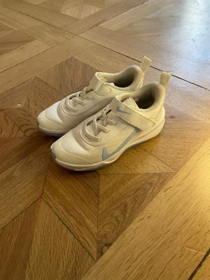 Vita Nike sneakers med grå detalj - Snygga vita Nike sneakers med ljusgrå swoosh och detaljer. Skorna har snörning och kardborreband för enkel påtagning. Ovandelen är i mesh och syntet för bra andningsförmåga och komfort. Perfekta för dig som gillar sportig stil och vill ha ett par lätta skor. Storlek 31