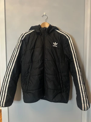Svart Adidas pufferjacka med huva - Svart pufferjacka från Adidas med ikoniska vita ränder längs ärmarna och Adidas-logga på bröstet. Jackan har huva, dragkedja framtill och två sidofickor. Perfekt för dig som gillar sportig stil och vill hålla dig varm under kyliga dagar.