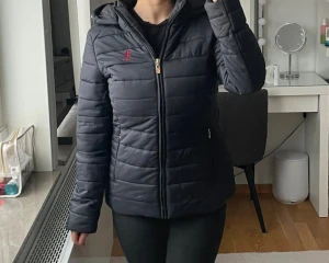 Svart pufferjacka från U.S. Polo Assn. - Svart pufferjacka med röd broderad logga på bröstet. Jackan har huva, guldfärgade dragkedjor och två sidofickor med dragkedja. Quiltad design och långärmad modell som ger en snygg och sportig look. Perfekt för kyliga dagar. Aldrig använd!