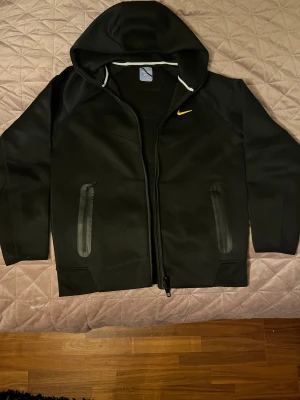 Svart Nike NOCTA vindjacka med huva - Svart vindjacka från Nike NOCTA med huva och gul logga på bröstet samt NOCTA-tryck på ärmen. Jackan har två fickor med dragkedja och stilrena sömmar. Materialet är mjukt och syntetiskt, perfekt för en sportig look.