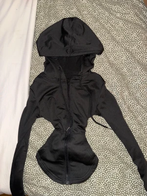 Svart croppad hoodie med dragkedja - Säljer en svart croppad hoodie med dragkedja framtill och huva. Tröjan har långa ärmar, figurnära passform och snörning i huvan. Perfekt till jeans eller träningsbyxor för en chill look.