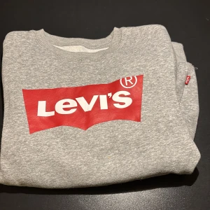 Grå sweatshirt från Levi's med röd logga - Säljer en grå sweatshirt från Levi's med den klassiska röda loggan tryckt framtill. Tröjan har rund hals och ribbade muddar vid ärmslut och nederkant. Perfekt för dig som gillar streetstyle och vill ha en enkel men snygg look.
