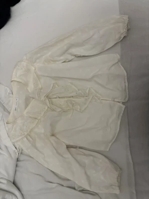 Vit blus med volang och spets från Zara - Romantisk vit blus från Zara med volangkrage och spetsdetaljer. Långa ärmar med resår vid handlederna och knappar framtill. Tunt, lätt material med diskret struktur och söt rosett vid halsen. I storlek 140.