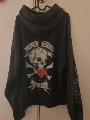 Svart Ed Hardy hoodie med dödskalle - Svart hoodie från Ed Hardy med stor färgglad dödskalle och texten 'Love Dies Hard' på ryggen. Framsidan har Ed Hardy-logga och ficka. Tryck även på ena ärmen. Mjuk bomullskänsla och klassisk huva med dragsko.