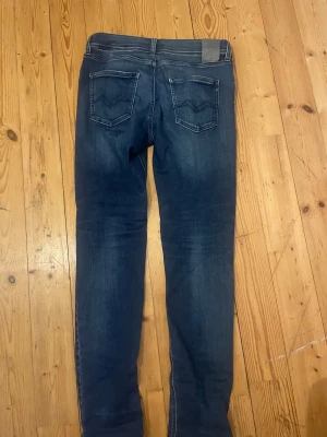 Replay blå jeans med raka ben - Snygga blå jeans från Replay med klassisk femficksdesign och raka ben. Jeansen har en liten slitning på höger knä samt en liten fläck, det är 100 cm långa, jag är 180 cm och väger 63 kg och de sitter lite tajt runt benen så ni bara vet.