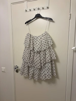 Prickig volangklänning från NA-KD - Superfin vit miniklänning från NA-KD med svarta prickar och flera lager volanger. Axelbandslös modell i luftig chiffong. Använd två gånger så som ny i skicket.