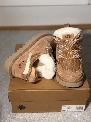 Uggs lowmel  - Snygga mörk beige brun sneakers från UGG med grov sula, snörning och insida i mjuk fårull. Ovandel i mocka och textil, detaljerade mönstrade skosnören och UGG-logga på sidan. Perfekta för kyliga dagar när du vill vara både varm och trendig.