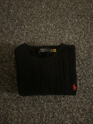 Svart kabelstickad tröja Polo Ralph Lauren - Svart kabelstickad tröja från Polo Ralph Lauren med rund hals och rött broderat logomärke på bröstet. Tröjan har ribbade muddar och är tillverkad i mjuk bomull, perfekt för dig som gillar klassisk stil med en modern touch.