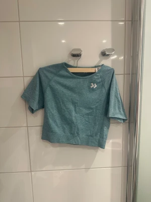 Blå croppad träningstopp från lager157 - Säljer en blå croppad träningstopp från lager157 med kort ärm och rund hals. Toppen har ventilerande meshdetaljer och en diskret logga framtill. Perfekt för gymmet eller löprundan.