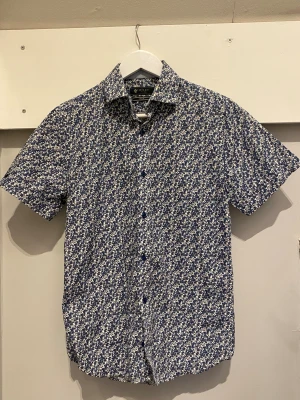 Blommig kortärmad skjorta från Riley - Kortärmad skjorta från Riley i slim fit med blommigt mönster i blått och vitt. Skjortan har klassisk krage, blå knappar och är gjord i mjuk bomull. Perfekt för sommarfesten eller under en mörkblå pullover