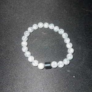 Vitt pärlarmband med marmormönster - Stilrent armband med vita pärlor och grått marmormönster, samt en större svart pärla som detalj. Elastiskt band för enkel användning. Perfekt accessoar för att lyfta din outfit.