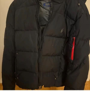 Svart pufferjacka Polo Ralph Lauren - Svart pufferjacka från Polo Ralph Lauren med broderad logga på bröstet och röd dragkedja på ärmen. Jackan har huva med snörning och klassisk quiltad design. Perfekt för kalla dagar och har en snygg, sportig look.