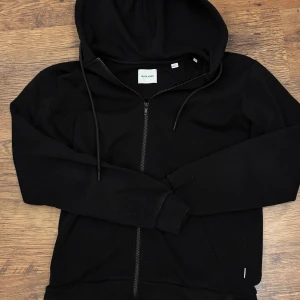 Svart hoodie från Jack & Jones - Svart ziphoodie från Jack & Jones med dragsko i huvan och två fickor framtill. Modellen är klassisk med ribbade muddar och mjukt sweat-material. Perfekt för chill dagar och enkel att matcha med jeans eller joggers.