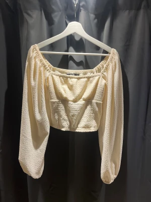 Offshoulder croppad topp från Bik Bok - Superfin offshoulder topp från Bik Bok i ljusbeige färg. Croppad modell med långa puffiga ärmar och smockad struktur i ett mjukt, stretchigt material. Perfekt för dig som gillar trendiga och feminina detaljer.