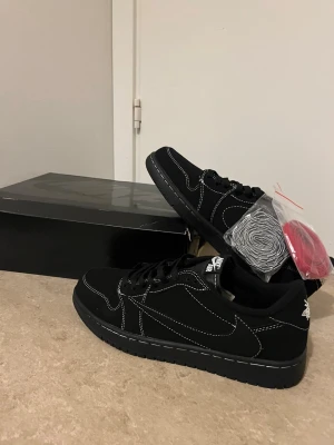 Svarta Nike Air Jordan 1 lows med broderi - Svarta Nike Air Jordan 1 lows med vit kontrastsöm och broderade detaljer på hälen, inklusive en bi och Air Jordan-logga. Skorna har platt sula, rund tå och kommer med extra röda skosnören. Materialet är mocka och syntet, och de har klassisk snörning.