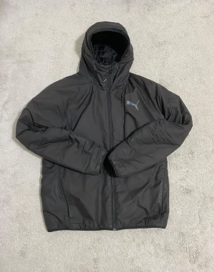Svart pufferjacka från Puma - Svart pufferjacka från Puma med huva och dragkedja framtill. Jackan har en stilren design med Puma-loggan i grått på bröstet. Perfekt för kalla dagar och har en bekväm passform med långa ärmar.