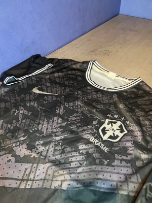 Brasilien fotbolls tröja  - Säljer en riktigt unik Brasilien-tröja i svart/grått med detaljerat camo-mönster. Tröjan har Nike-loggan och Brasilien-emblemet tryckt på bröstet, samt snygga vita detaljer runt kragen och ärmarna. Materialet är lätt, bekvämt och andas bra – perfekt för både träning och vardag.