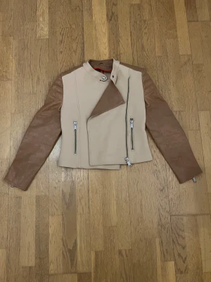 J Lindeberg läderjacka - strl.36 - Snygg bikerjacka i beige med bruna läderärmar och coola quiltade detaljer på ryggen. Jackan har asymmetrisk dragkedja framtill, två fickor med dragkedja och ståkrage med knapp. Insidan har röda ränder för extra stilpoäng. Perfekt för dig som gillar edgy och modern look. 