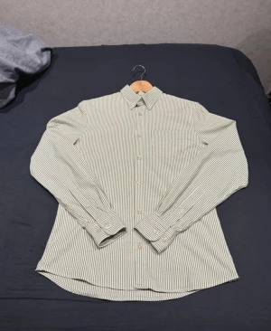 Randig skjorta från J.Lindeberg - Snygg slim fit skjorta från J.Lindeberg i storlek S. Skjortan har ett klassiskt randigt mönster i ljusgrönt och vitt, knappar framtill och en bröstficka. Tillverkad i mjuk bomull och har långa ärmar med manschettknappar. Perfekt för dig som gillar stilrena och fräscha plagg.