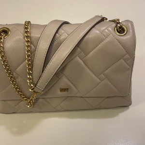 Ljus grå quiltad axelväska från DKNY - Snygg ljus grå axelväska från DKNY i quiltat skinn med guldfärgade kedjedetaljer. Väskan har ett praktiskt innerfack med dragkedja och DKNY-logga på framsidan. Perfekt storlek för det viktigaste och stilren design som lyfter din look. Ovbservera. Väskan har små skrapmärken på baksidan till höger. 