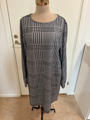 Rutig långärmad klänning Jacqueline de Yong - Snygg långärmad klänning från Jacqueline de Yong i svartvitt rutigt mönster. Modellen är rak och knälång med rund halsringning. Perfekt för dig som gillar klassiska och stilrena plagg med en modern twist.