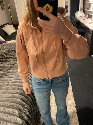 Persikofärgad hoodie från lager 157 - Säljer en persikofärgad hoodie från lager 157 i storlek M. Tröjan har huva med snörning, dragkedja framtill och ribbade muddar vid ärmslut och nederkant. Materialet är mjukt och känns som bomull, perfekt för chill dagar. Passformen är loose och den är långärmad.