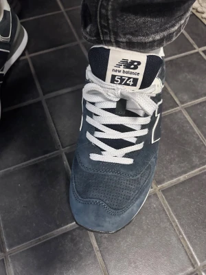 New Balance 574 blå sneakers - Säljer ett par New Balance 574 sneakers i mörkblå mocka med vita detaljer och vit sula. Skorna har klassisk N-logga på sidan, vit snörning och meshpartier för extra komfort. Perfekta för dig som vill ha en clean och sportig look.