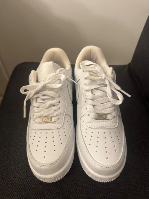 Nike Air Force 1 vita sneakers - Klassiska Nike Air Force 1 sneakers i helvitt skinn med perforerad tå, rund tå och vit sula. Skorna har snörning och den ikoniska swoosh-loggan på sidan. Perfekta för dig som vill ha en clean och tidlös look.