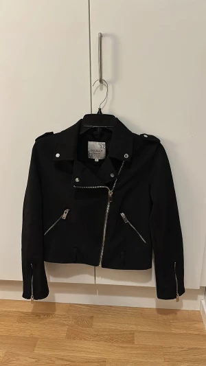 Svart skinnjacka Orcelly Fashion S - Snygg svart skinnjacka från Orcelly Fashion i storlek S. Jackan har klassisk bikerstil med silvriga dragkedjor, knappar och axelklaffar. Croppad passform och långärmad design, perfekt för dig som gillar en edgy look.