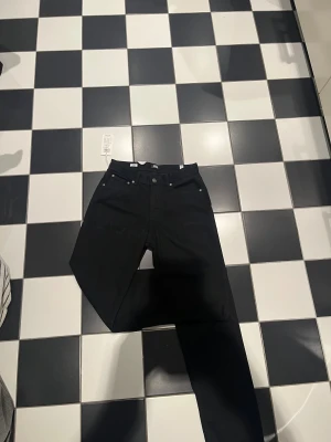 Svarta jeans från Black Jones, 164 - Svarta jeans från Black Jones i rak modell med klassisk femficksdesign och knappstängning. Jeansen har normal passform och är tillverkade i ett slitstarkt bomullsmaterial. Perfekt för dig som gillar stilrena och enkla svarta jeans. Perfekt julkalapp nu till vintern, jeansen är alltid använda och alla prislappar och sånt är kvar!
