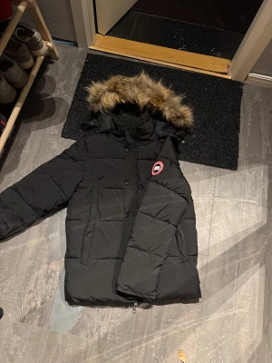 Canada goose jacka - Säljer en svart dunjacka från Canada Goose med pälskant på huvan och klassisk logga på ärmen. Jackan har dragkedja och knappar framtill, stora fickor och quiltad design. Perfekt för kalla vinterdagar och har en snygg, lite oversized passform.