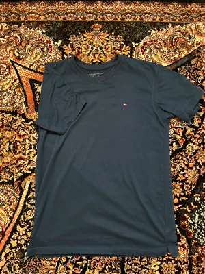 Mörkblå t-shirt från Tommy Hilfiger - Snygg mörkblå t-shirt från Tommy Hilfiger med klassisk rund hals och liten broderad logga på bröstet. Modellen är enkel och stilren, perfekt för dig som gillar cleana plagg. Tillverkad i mjuk bomull som känns skön mot huden.