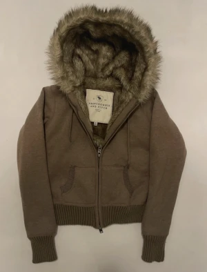 Brun hoodie med fuskpäls från Abercrombie - Snygg brun hoodie från Abercrombie & Fitch med dragkedja, två fickor och stor huva fodrad med mjuk fuskpäls. Ribbstickade muddar och midja ger en schysst passform. Perfekt för dig som gillar en mysig och trendig look. Skickas inom 24 timmar 