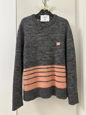Grå stickad tröja Acne Studios - Snygg gråmelerad stickad tröja från Acne Studios med rosa och mörkgrå ränder nedtill. Klassisk rund hals och långärmad modell. På bröstet sitter Acne Studios ikoniska rosa ansiktsmärke. Perfekt för dig som gillar stilrena detaljer och skandinavisk design.